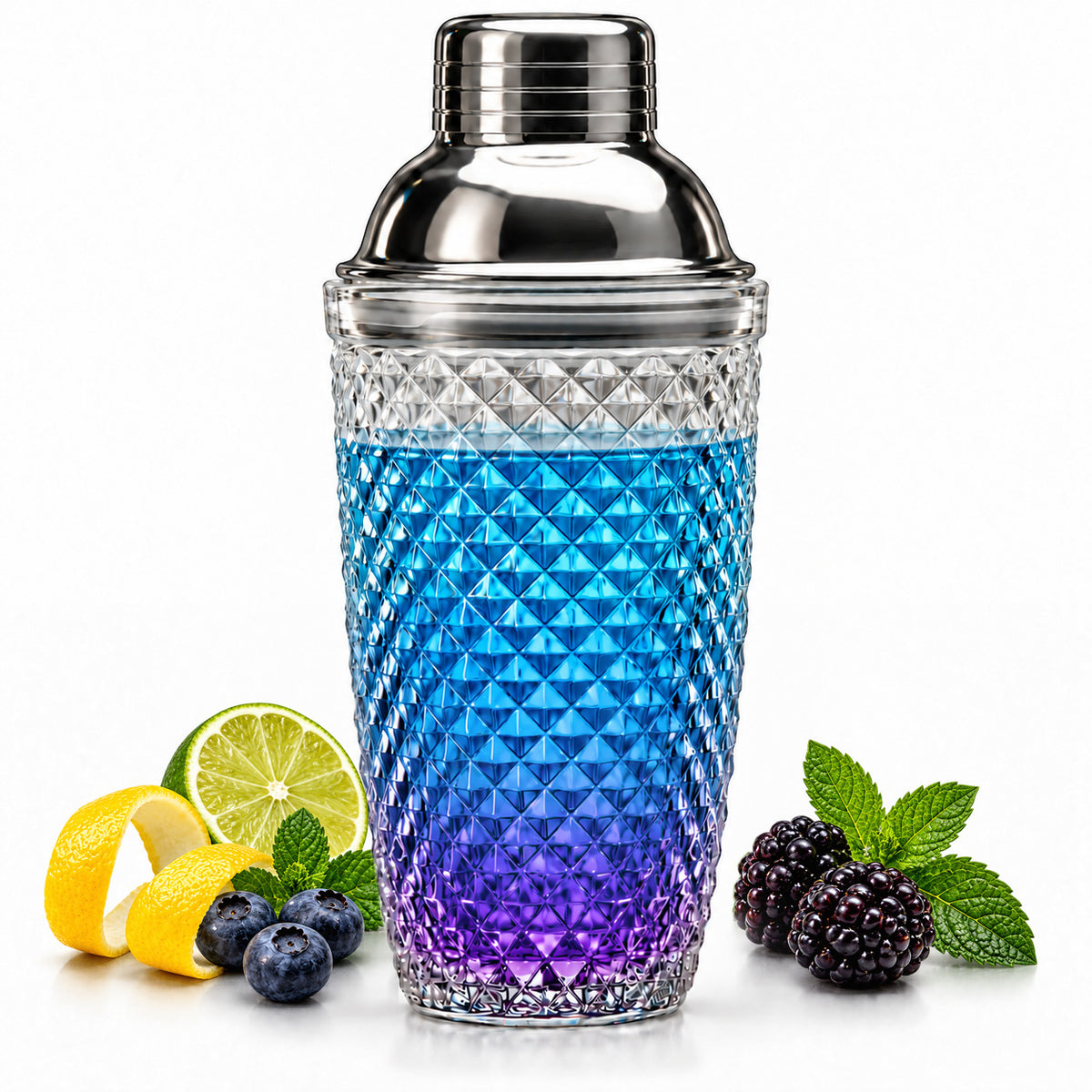 Diamond cut glass cocktail shaker with a blue and purple drink, lime, lemon peel, berries and mint